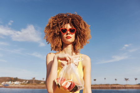Young woman with curly hair holding a bag of fruit on a sunny beachの写真素材