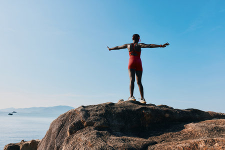A person standing on a rock by the sea with arms outstretched, embracing the freedom and beauty of nature against a clear blue skyの写真素材