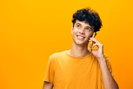 young man talking on a yellow phone, smiling happily, wearing a casual orange t shirt against a vibrant yellow background, expressing joy and connection with technology or friendsの写真素材