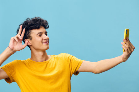 Young man taking a selfie with a smartphone against a blue background, smiling and making a peace sign with his hand in a casual and joyful poseの写真素材