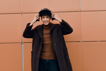 Fashionable young man in brown coat standing in front of orange wall with hands on head and wearing...
