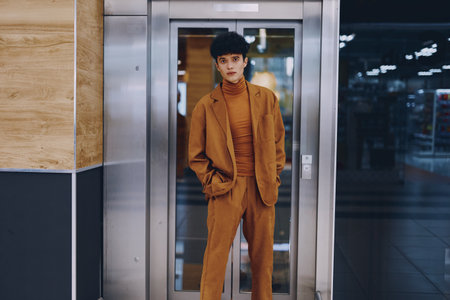 Fashionable young adult in a stylish brown suit standing in front of an elevator in a modern buildingの写真素材
