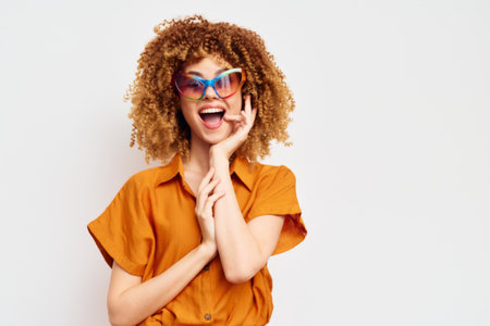 Happy young woman with curly hair wearing colorful sunglasses, expressing joy against a light background, perfect for summer fashion themes and lifestyle imageryの写真素材
