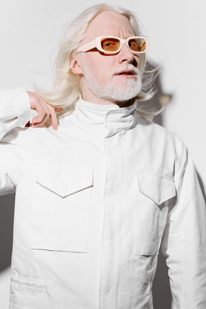 Fashionable elderly man wearing stylish white jacket and sunglasses against a minimalist white background, exuding confidence and contemporary style in modern aestheticsの写真素材