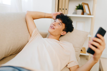 Relaxed young man lounging on a couch with a smartphone, smiling while looking up in a bright, cozy room with bookshelves Natural light creating a warm atmosphereの写真素材