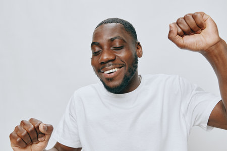 Happy young man celebrating joyfully in a white t shirt on a light background, expressing excitement with arms raised and a broad smile, showcasing positive emotionsの写真素材