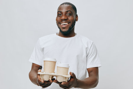 Smiling man holding coffee cups in a takeaway holder, cheerful expression, casual white t shirt,...