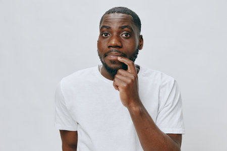 Thoughtful man in a white t shirt, looking contemplative with a finger to his lips, neutral background, expressing curiosity and reflection in a clean settingの写真素材
