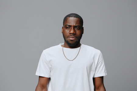 Male model with a serious expression wearing a plain white t shirt, standing against a neutral grey...
