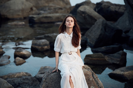 Serene woman in white dress sitting on rocky shore overlooking ocean wavesの写真素材