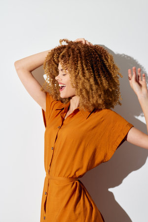 Woman with curly hair in a stylish orange dress, celebrating against a minimalist white background The image captures joy and elegance beautifullyの写真素材