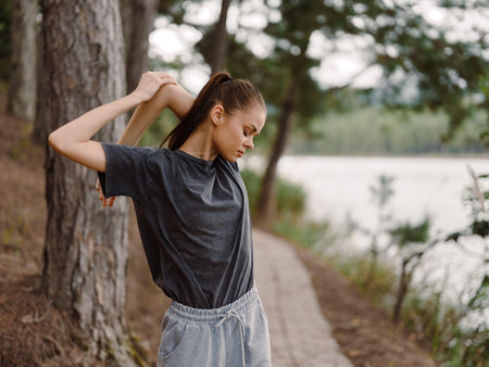 Young woman stretching outdoors by the lakeside, wearing a casual grey t shirt and sweatpants, enjoying nature and personal wellness Fitness and health conceptの写真素材