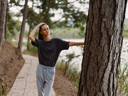 Thoughtful young woman in casual gray outfit enjoying nature by the lake, walking along the pathway, surrounded by trees and greenery Serenity and mindfulness conceptの写真素材