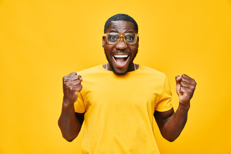 Happy African man celebrating success with enthusiasm in a bright yellow shirt against a vibrant yellow background, expressing joy and excitementの写真素材