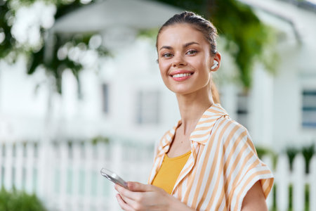 Smiling woman using smartphone outdoors with wireless earbuds, wearing a striped shirt, in a lush green setting with soft sunlight highlighting her featuresの写真素材
