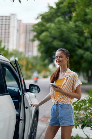 Young woman standing next to a car, smiling, wearing casual summer clothing with a phone in hand, surrounded by greenery and urban buildings in the backgroundの写真素材