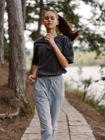 Young woman jogging along a natural pathway by the lake, dressed in casual sportswear and enjoying...