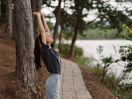 Relaxed young woman stretching outdoors by the lake, enjoying nature and fresh air, with trees and water creating a serene atmosphere Wellness and rejuvenation themeの写真素材