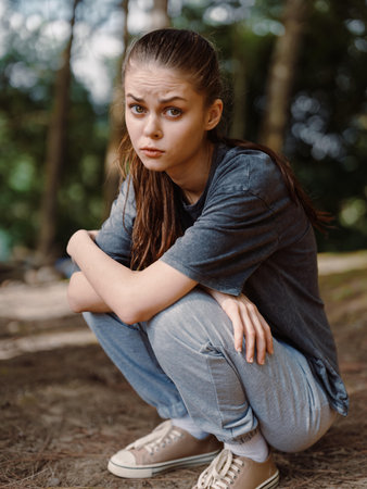 Pensive young woman sitting on forest floor, wearing casual outfit, reflecting in nature, with trees and greenery surrounding her in a tranquil settingの写真素材