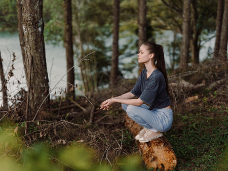Calm young woman sitting in nature, meditating on a log by a serene body of water Peaceful and tranquil atmosphere surrounded by trees Mindfulness and relaxation conceptの写真素材