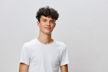 Young smiling man with curly hair in a plain white t shirt, standing against a light gray...