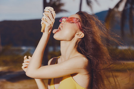 Young woman enjoying ice cream on a sunny beach with palm trees in the background She wears pink sunglasses and licks the ice cream cone with enthusiasmの写真素材