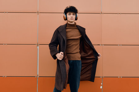 Young man in brown coat standing against orange wall with hands in pockets and headphones, smilingの写真素材
