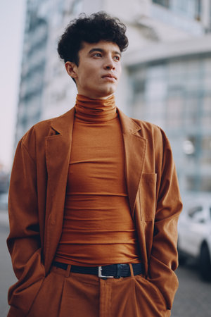 Young man in a stylish brown suit with a high neck top, standing outdoors, exuding confidence against a modern urban backdropの写真素材