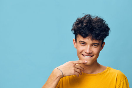 Young man with curly hair smiling playfully against a bright blue background He wears a yellow t...