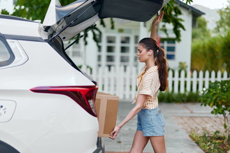 Young woman unloading boxes from a car in a residential setting, surrounded by greenery and a white picket fence Bright and sunny day with casual attireの写真素材