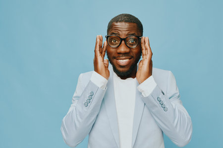 Happy man wearing glasses and light blue suit smiling with hands on face, expressing surprise or joy against a solid light blue background Energetic and vibrant conceptの写真素材