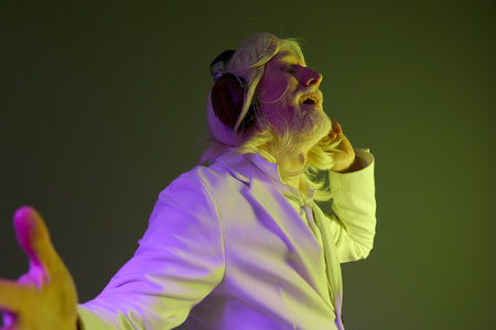 Stylish elderly man with long hair in a white suit enjoying music with headphones against a soft...