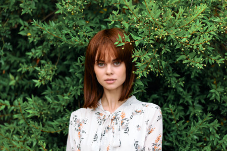 Vibrant redhead woman standing confidently in front of lush green bush with vibrant leaves