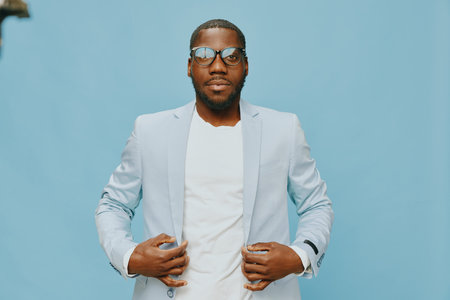 Stylish man in light blue suit, with glasses, confidently posing against a solid blue background, showcasing elegance and modern fashion for professional settingsの写真素材