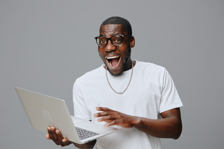 Young man expressing excitement while using a laptop, wearing casual white t shirt and glasses, neutral gray background, showcases happiness and enthusiasm in modern lifestyleの写真素材