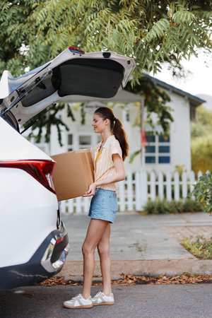 Delivery, lifestyle, woman, car, package, summer, outdoor, cheerful, casual, unloading, transportation, home, environment, nature, smiling, young A young woman is unloading a cardboard box from the back of a car, enjoying a sunny day outdoors near a houseの写真素材