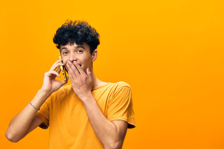 Surprised young man talking on a phone, wearing a bright yellow t shirt, with an expressive facial expression, against a vibrant orange background Engaging and lively conceptの写真素材
