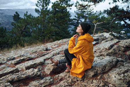 A woman in a yellow raincoat sits serenely on a rocky outcrop surrounded by majestic mountain peaksの写真素材