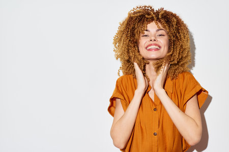 Happy young woman with curly hair smiling and touching her face, wearing a brown shirt against a...