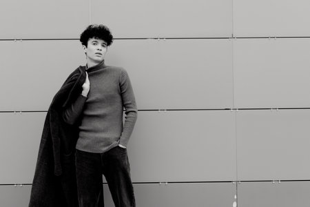 Fashion photo of a man in black and white, leaning against a wall with a coat draped over his shoulderの写真素材
