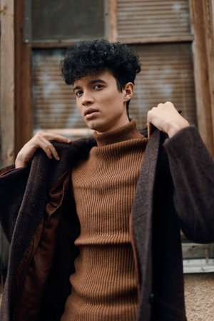 Stylish young man with curly hair in a brown sweater and coat standing by a window with hands on hipsの写真素材