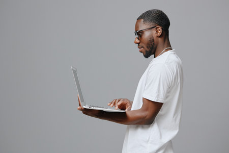 Young man using laptop, casual white t shirt, glasses, gray background, focused expression, technology concept Emphasizing modern lifestyle and connectivityの写真素材