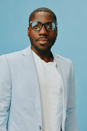 Stylish African man wearing glasses and a light blue blazer, posing confidently on a solid blue...