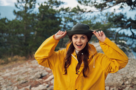 A woman in a yellow raincoat and hat standing on a rocky hill with hands on head Exploring the beauty of nature under the yellow rainの写真素材
