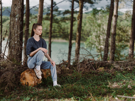 Thoughtful young woman sitting on a log in a tranquil forest setting, surrounded by trees and greenery, reflecting on nature and lifeの写真素材