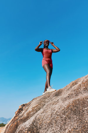 Confident woman standing on a rock under a blue sky, wearing a stylish outfit and sunglasses, exuding a sense of freedom and adventureの写真素材