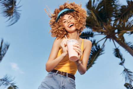 Cheerful young woman enjoying a smoothie outdoors against a clear blue sky and palm treesの写真素材