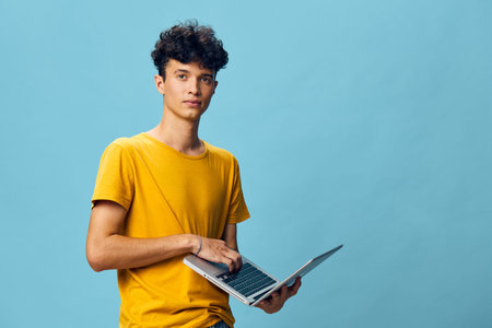 Young man with curly hair using a laptop against a light blue background, wearing a bright yellow t...