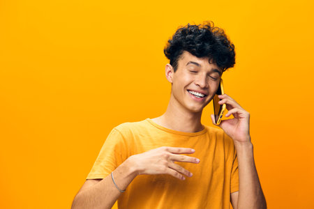 young man talking on a smartphone, smiling, yellow background, bright colors, casual attire, joy, communication, happiness, modern lifestyle, cheerful expression, contemporary portraitの写真素材
