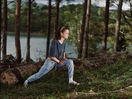 Focused young woman stretching outdoors in a natural setting, wearing casual athletic clothing near...